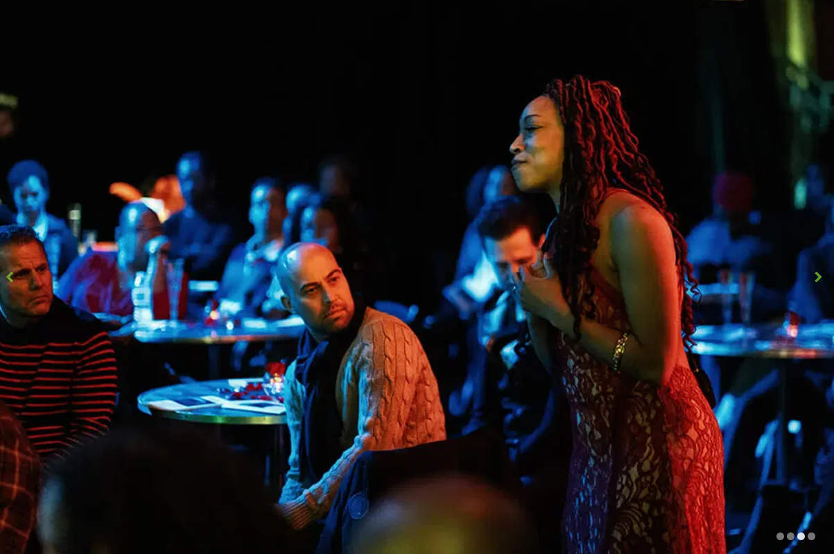 Performer in a lace gown lit in red among a blue-lit cabaret audience