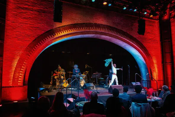 Wide shot of the stage under a red-lit arch with band and dancer