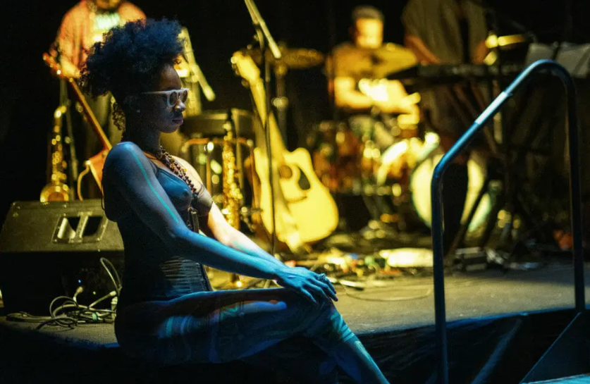 Performer seated at the edge of the stage in front of the band
